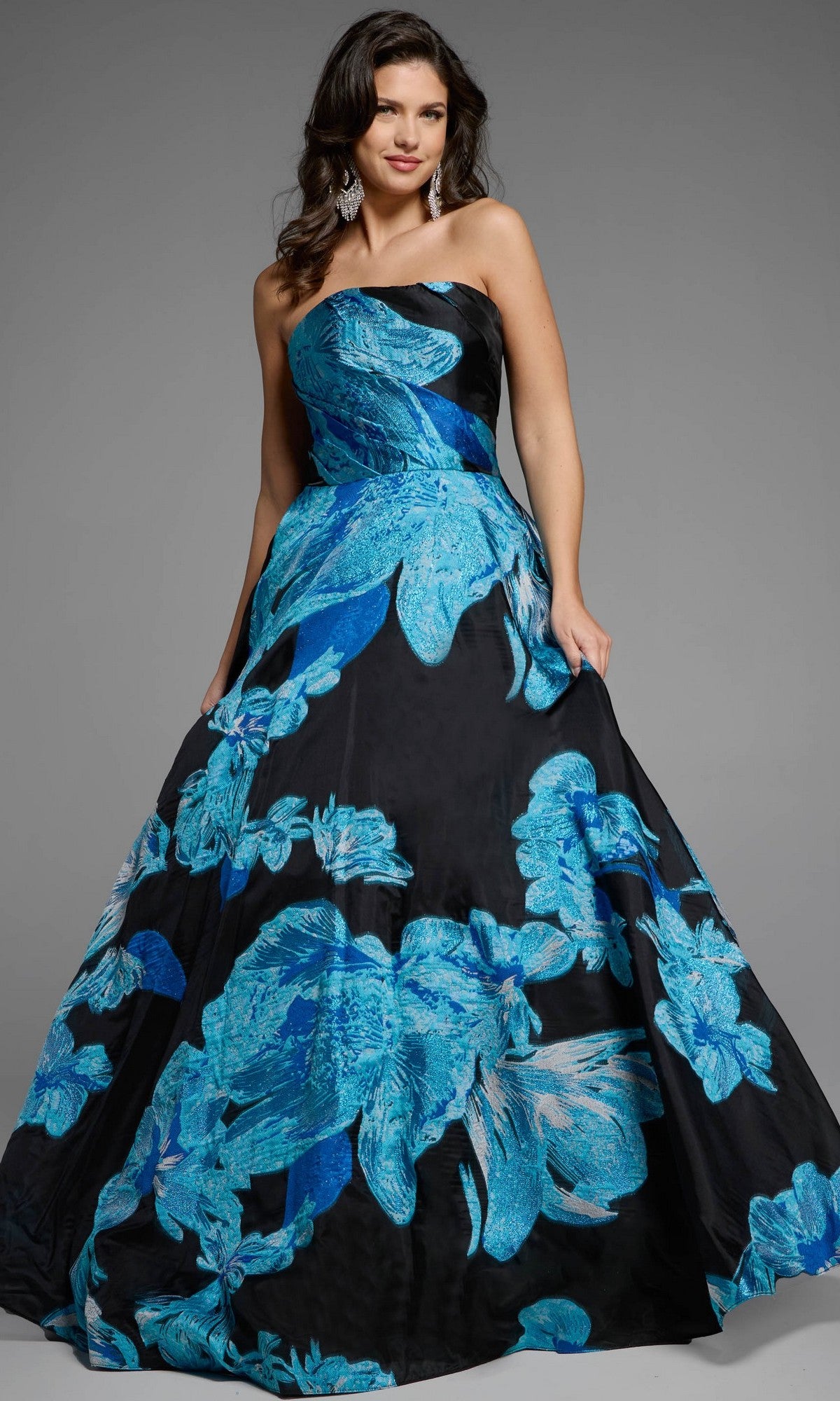 A woman models the Jovani 42436 long prom dress in blue/black, a strapless floral ball gown with bold blue patterns, standing against a plain gray background to highlight its elegance.